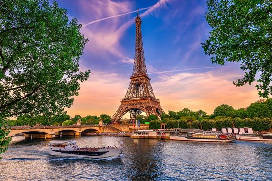 The Eiffel Tower with a vibrant sunset in the background and a body of water in the foreground with a boat passing in front