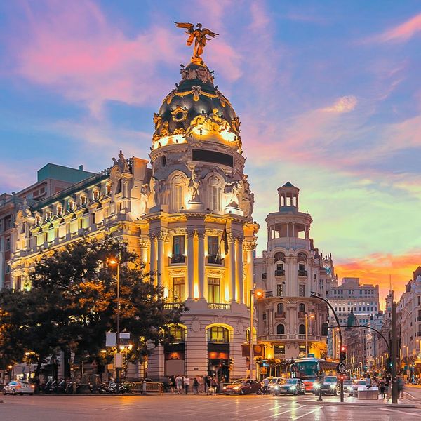 A bustling city street with ornately detailed buildings lit up at night with a pink sunset in the background