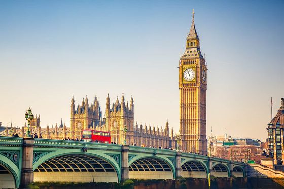A long green bridge with a red bus traveling on top of it and many tall ornate gold buildings in the background
