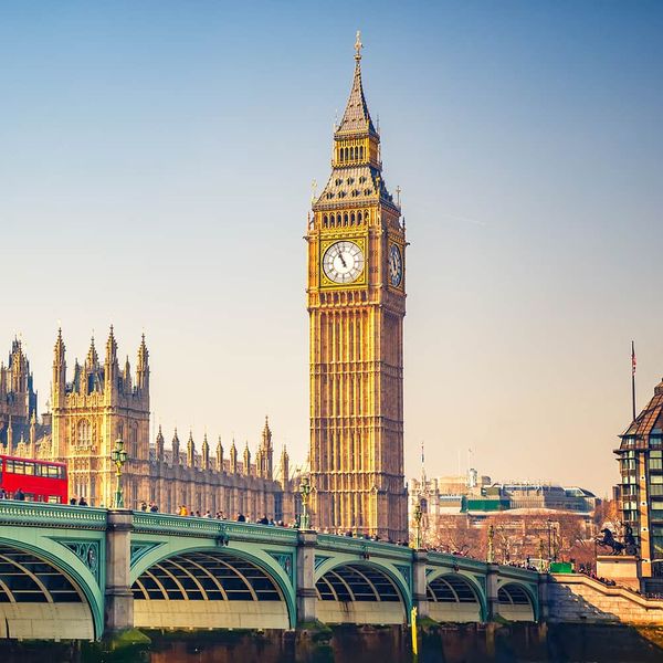 A long green bridge with a red bus traveling on top of it and many tall ornate gold buildings in the background