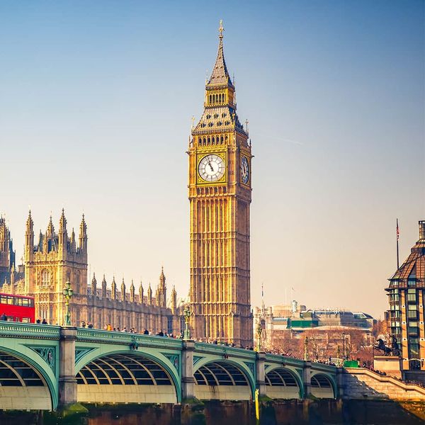 A long green bridge with a red bus traveling on top of it and many tall ornate gold buildings in the background