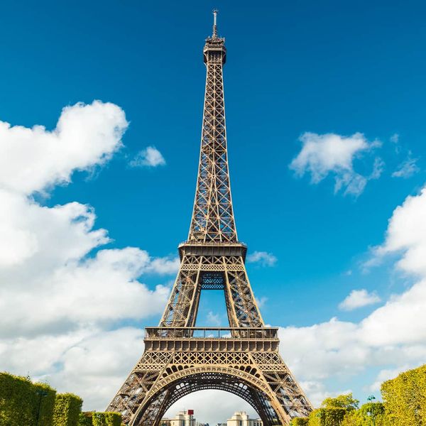 The Eiffel Tower in front of a blue sky with many white clouds scattering the sky and manicured trees lining the greenway in front of the Eiffel Tower