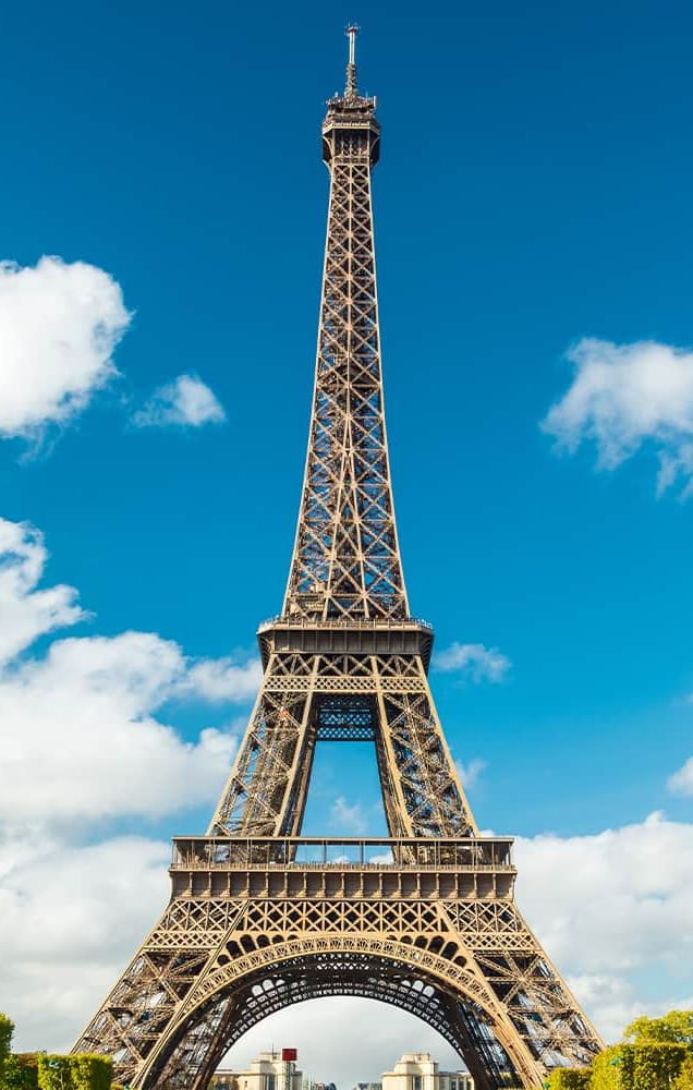 The Eiffel Tower in front of a blue sky with many white clouds scattering the sky and manicured trees lining the greenway in front of the Eiffel Tower