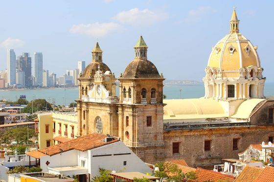 Aerial view of an ornate yellow building with three domed towers with ocean and a modern cityscape in the background