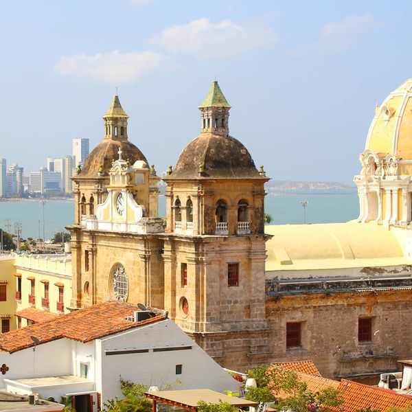 Aerial view of an ornate yellow building with three domed towers with ocean and a modern cityscape in the background
