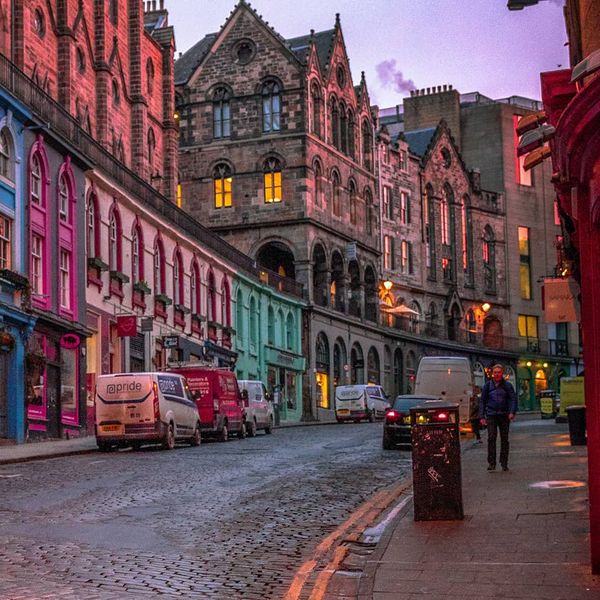 A row of vibrantly colored homes next to a street with cars parked along the side and people walking on the sidewalk