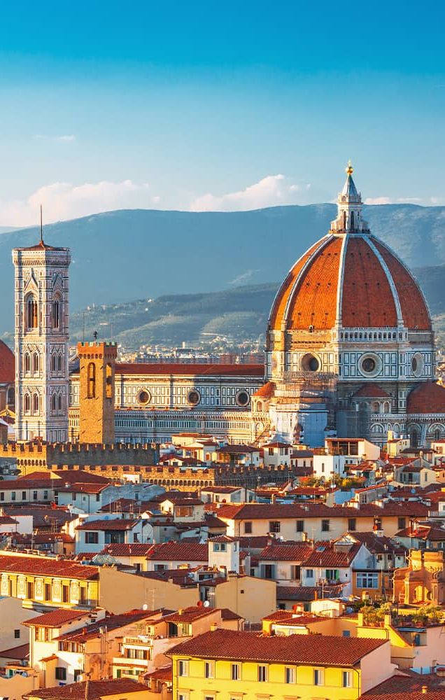 A view of the city of Florence with large red-domed buildings and a mountain range in the background 