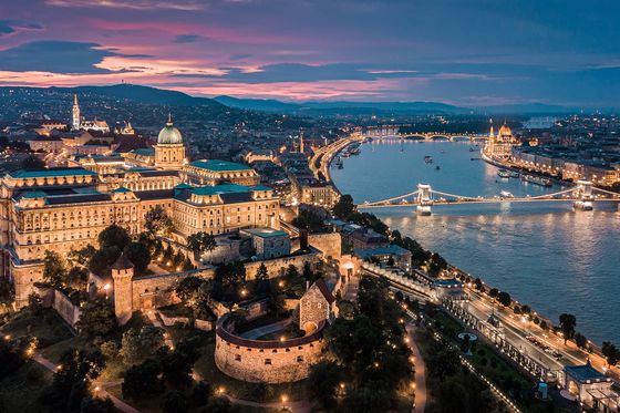 A large city with a grand central building situated next to a river with several bridges and everything is lit up with lights during dusk