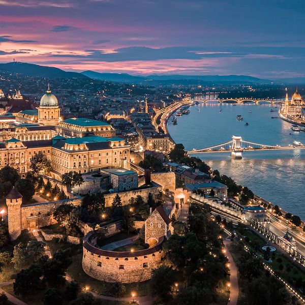 A large city with a grand central building situated next to a river with several bridges and everything is lit up with lights during dusk