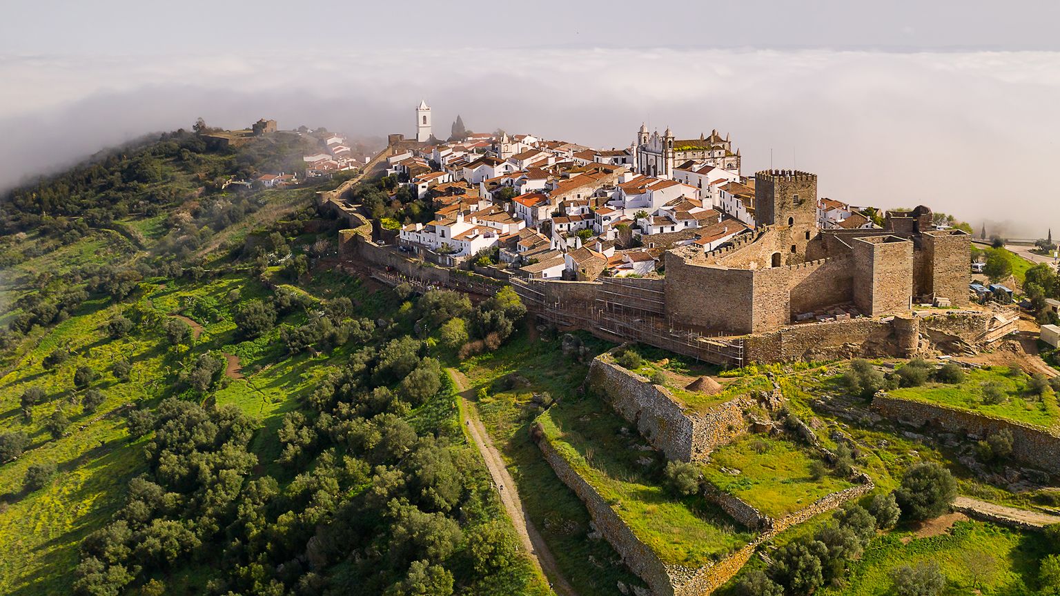 An establishing shot of Monsaraz village atop green mountains in the bottom left of the photo and foggy clouds in the top right of the photo.
