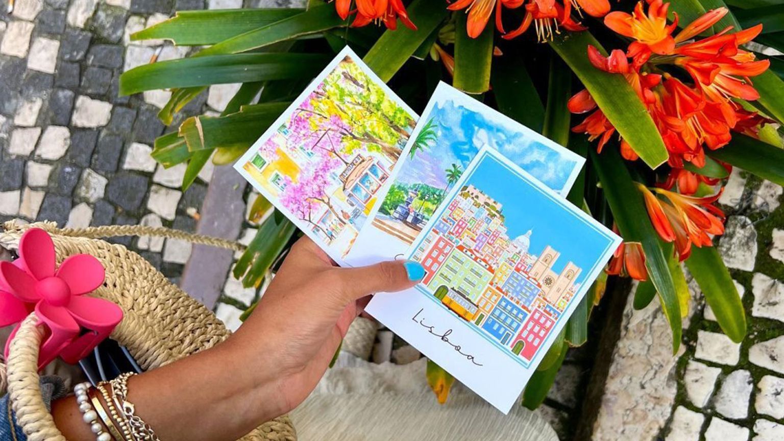 Photo taken of a hand holding 3 colorful Lisbon postcards, hovering over a vibrant orange flower bush.