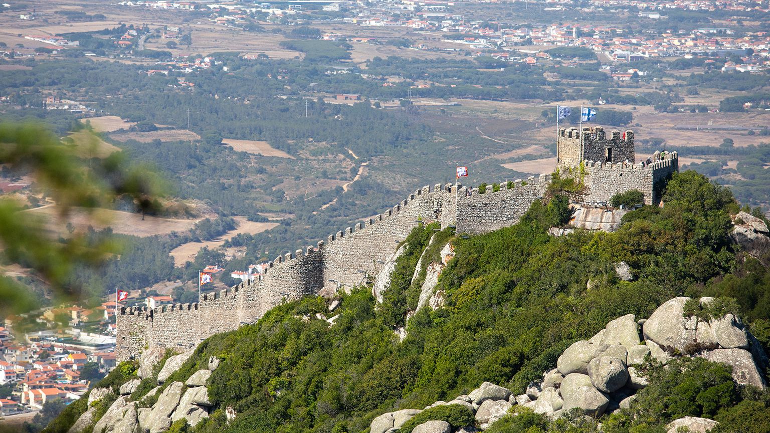 An establishing shot of part of Sintra castle in the middle of the photo. Green, rocky, mountains in the bottom of the photo and nearby villages out of focus in the background.