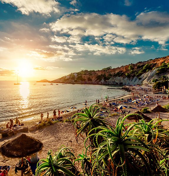 The shoreline of a calm beach during sunset with many people scattered around the sand and seaside cliffs in the background