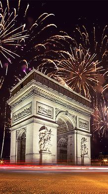 An extremely large beige stone arch with many detailed and symmetrical carvings all over the arch and fireworks lighting up the night sky in the background