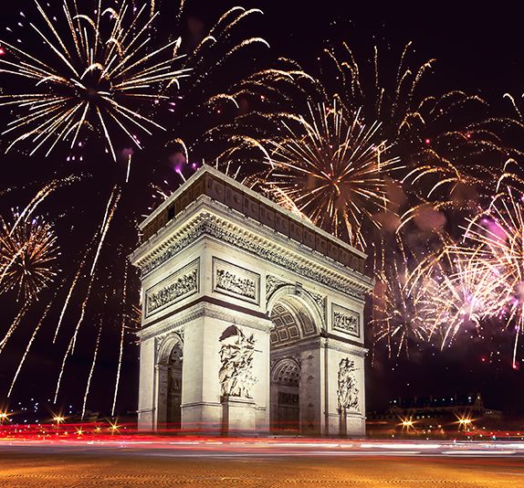 An extremely large beige stone arch with many detailed and symmetrical carvings all over the arch and fireworks lighting up the night sky in the background