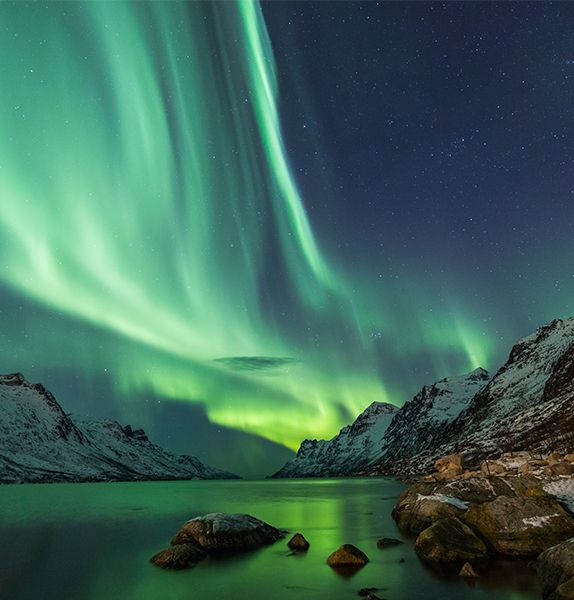 A frozen lake with rocky snow-covered mountains surrounding it and bright green Northern lights shining through the night sky and reflecting off the frozen lake
