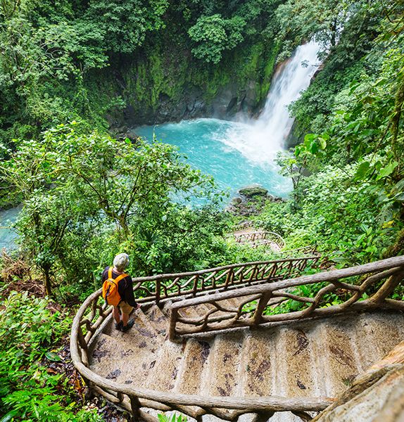 A person with a backpack standing in the middle of a staircase located in the middle of a lush green jungle and a gushing waterfall in the background