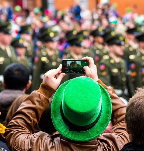 A person in a bright green hat taking a picture on his cellphone of a group of people walking in a parade
