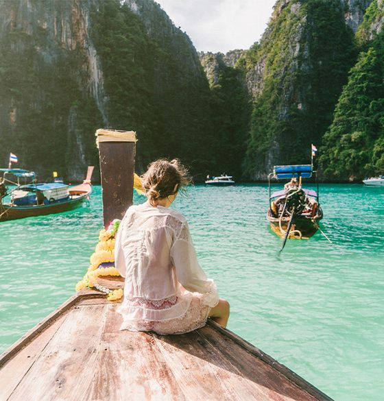 A woman in a white dress sitting at the edge of a boat with her feat touching the clear blue water and foliage-covered cliffs in the background