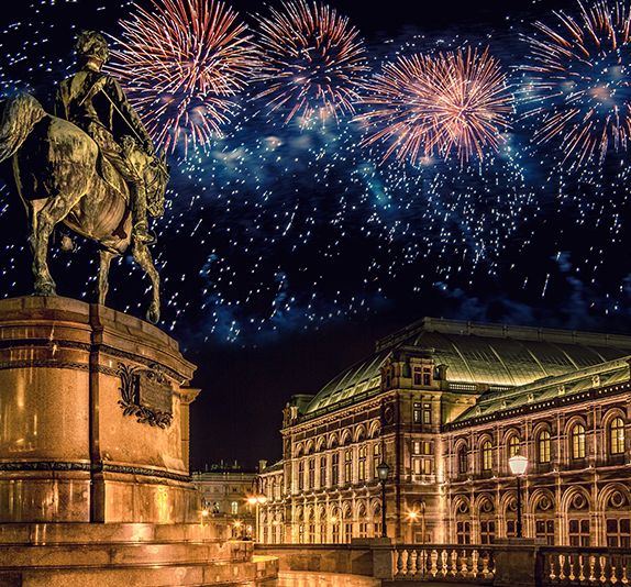 A grand statue of a figure riding on a horse nearby a huge ornate mansion during nighttime with fireworks covering the night sky