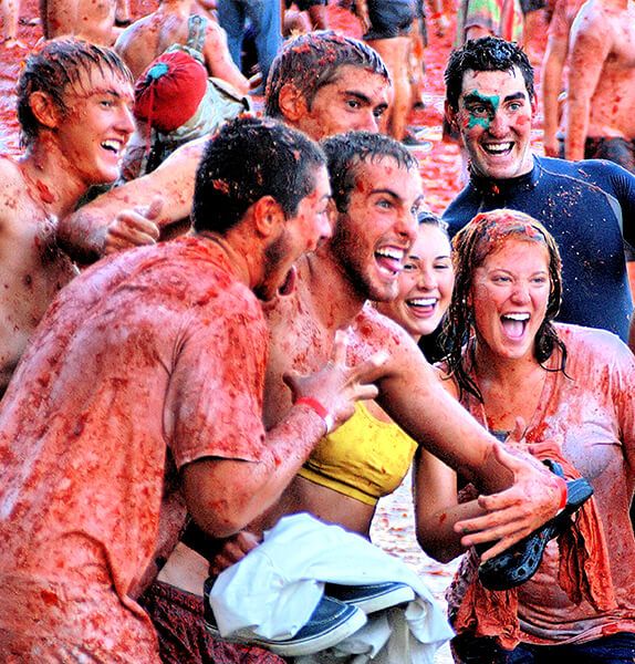 Several people with their mouths open and teeth showing in an excited expression covered from head to toe in the red juices of tomatoes