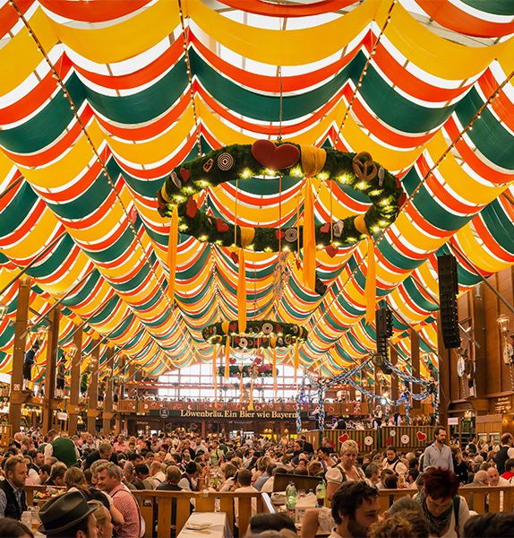 An outdoor hall filled with many people sitting down at tables with green, red, and yellow ribbons decorating the top of the hall