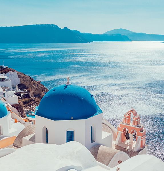 White buildings with a bright blue dome on top situated at the edge of a hill overlooking the calm ocean with other islands in the background