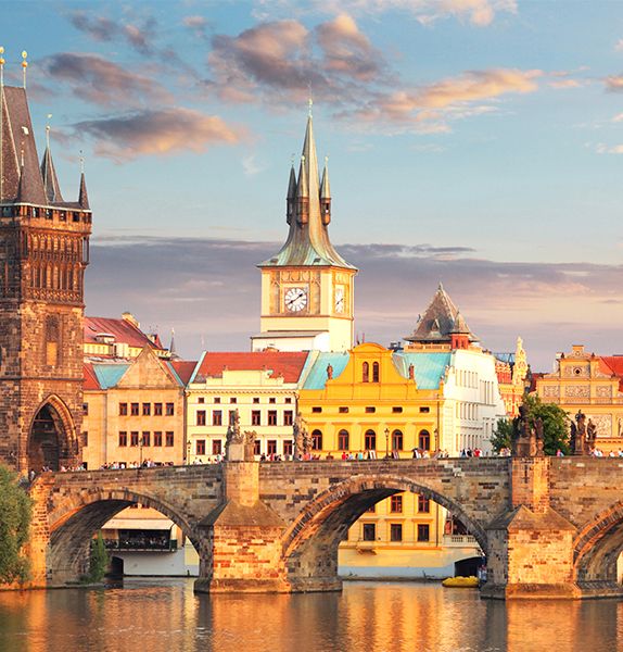 A view of a stone bridge during sunset with a clock tower and bright vibrant buildings in the background