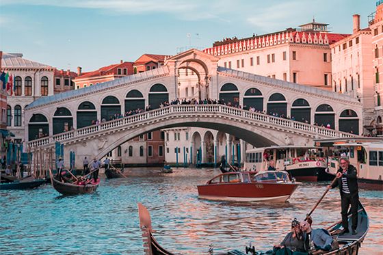 Several people traveling through water on boats with a pedestrian bridge in the background and buildings surrounding the water