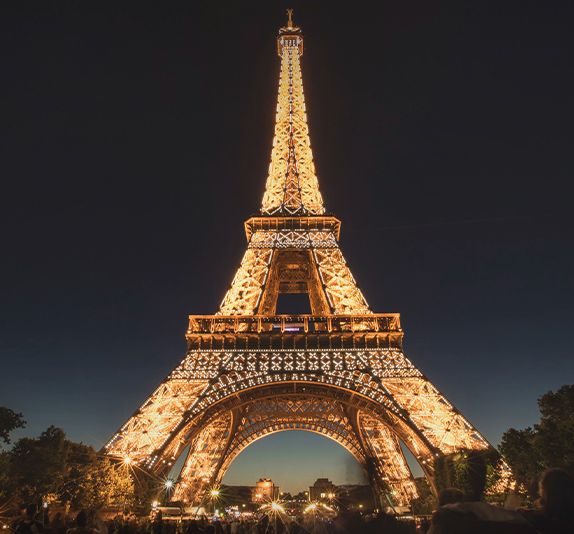 A bottom-up view of the Eiffel Tower lit up against the dark night sky with several people standing around the base of the tower