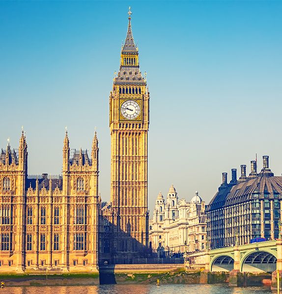 An extremely tall clock tower with many ornate details surrounded by several other grand buildings with a blue sky in the background