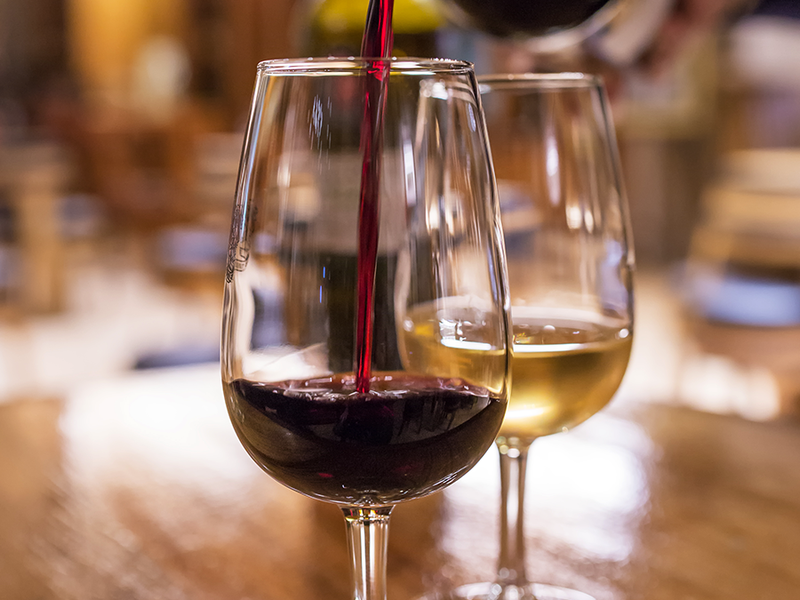 Two wine glasses on a wooden table, one being filled with red wine from a bottle and the other containing white wine, with a blurred indoor background suggesting a restaurant or bar setting.