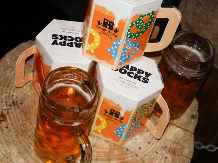 Ein Happy Socks-Geschenkset in Form eines Bierkrugs, und neben dem Krug stehen drei Happy Socks mit Oktoberfest-Motiven.