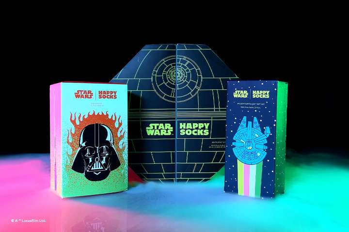 3 Star Wars x Happy sokken Geschenkdozen die uit een donkere omgeving tevoorschijn komen, omgeven door witte rook die wordt geaccentueerd met neonkleuren.