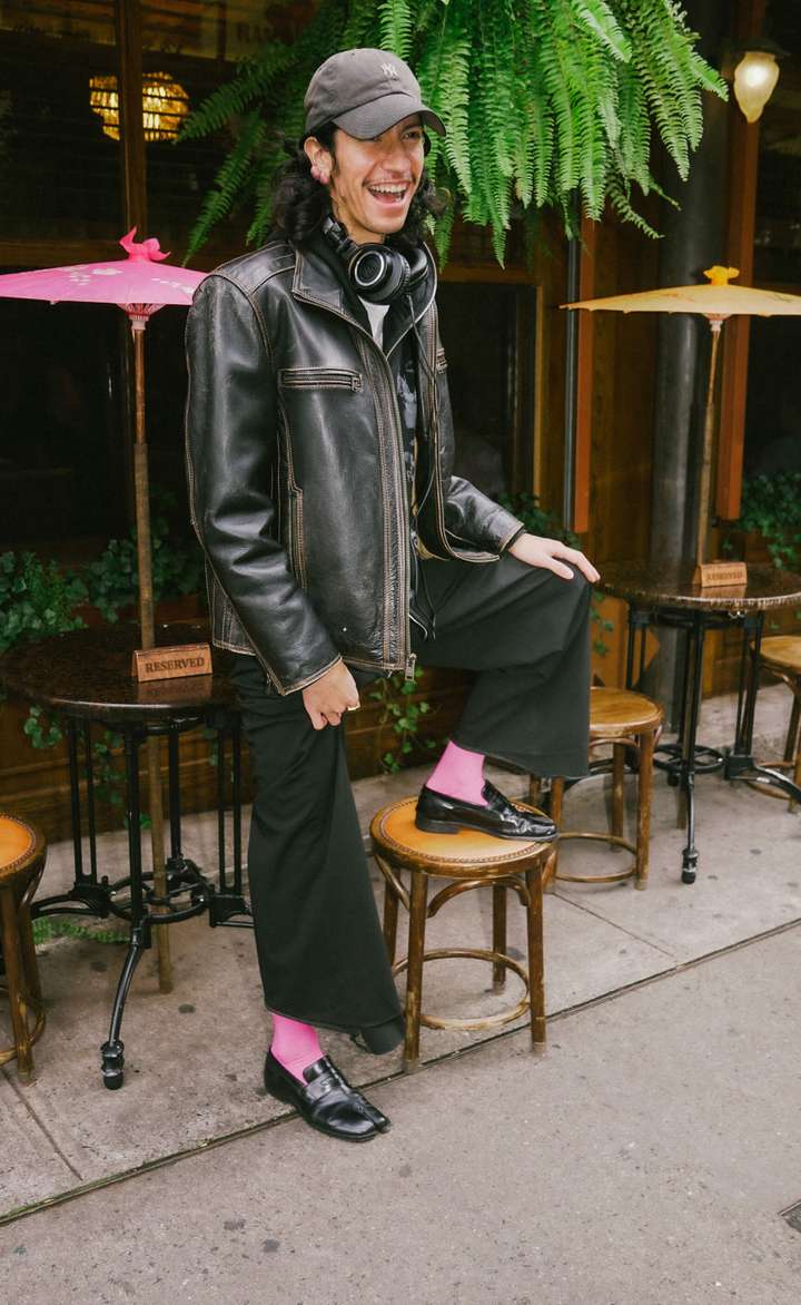 A man standing outside a bar, wearing a full black outfit with a cap, oversized leather jacket, flared pants, shiny loafers and pink happy socks solid socks.