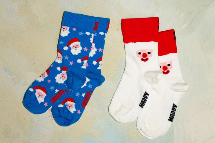  Ein Bild, das ein Paar Kinderfüße zeigt, die braune und grüne Happy-Socks-Kindersocken mit Weihnachtsbaummotiv tragen