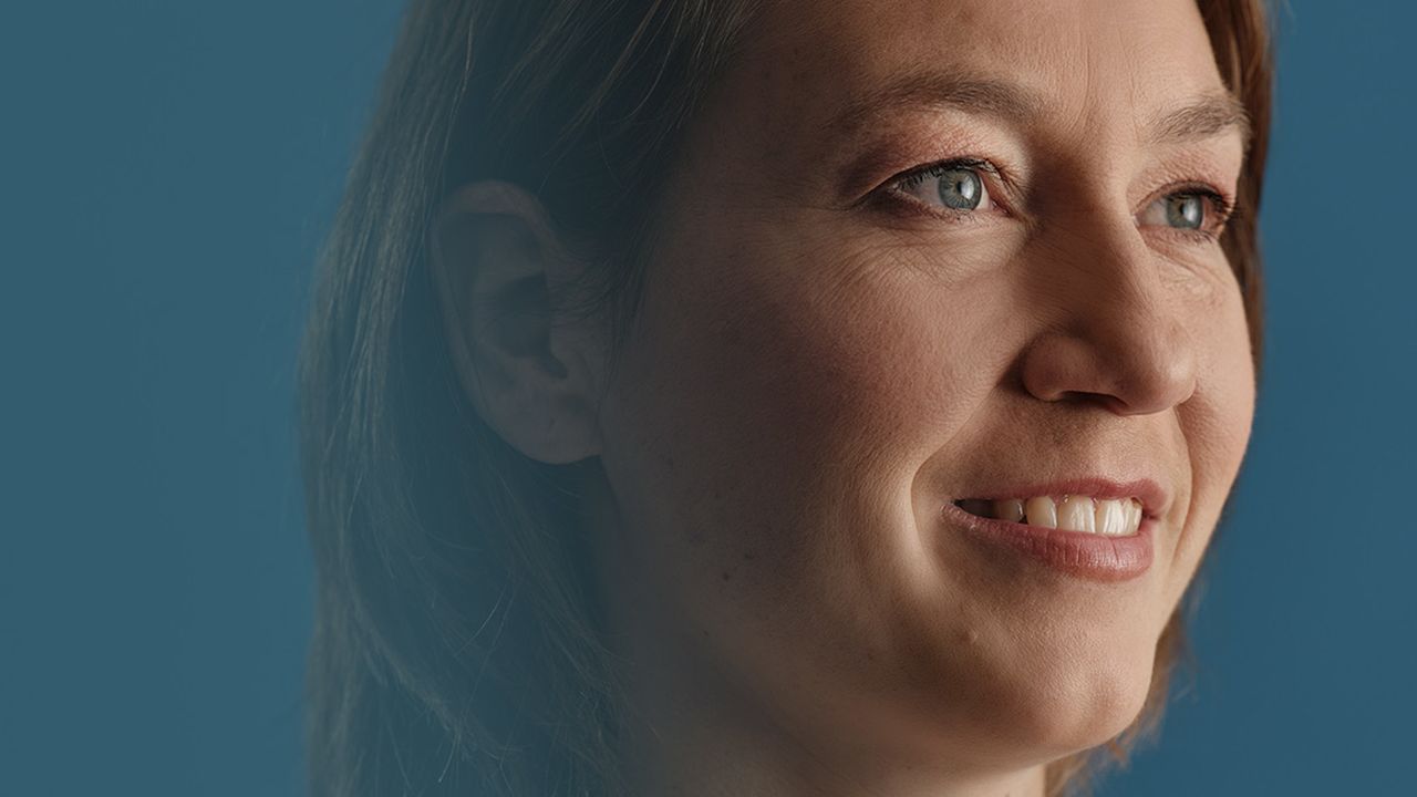 A close-up of a smiling woman with brown hair, looking sideways against a solid blue background.