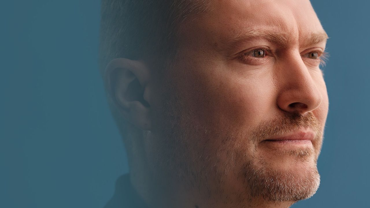 Profil d'un homme aux cheveux courts et à la barbe de trois jours, regardant pensivement au loin sur fond bleu.