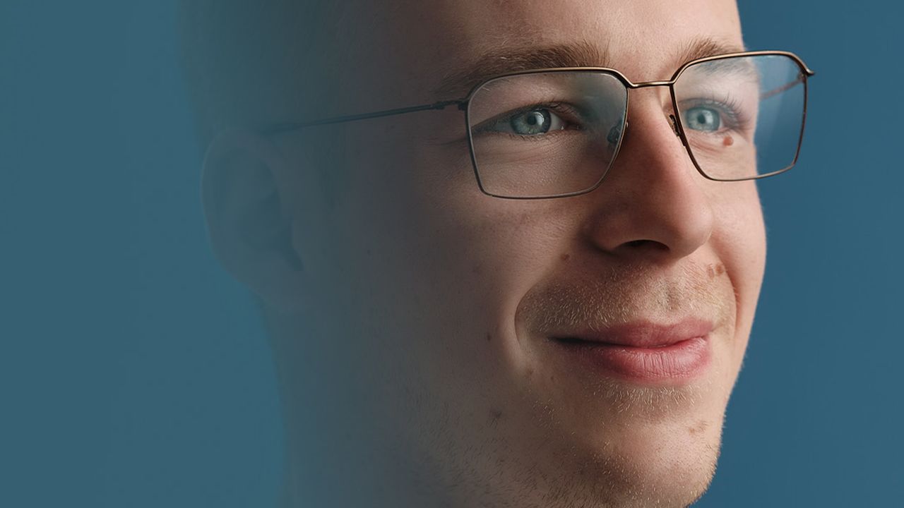 Man with glasses smiling gently, gazing upward against a blue background.