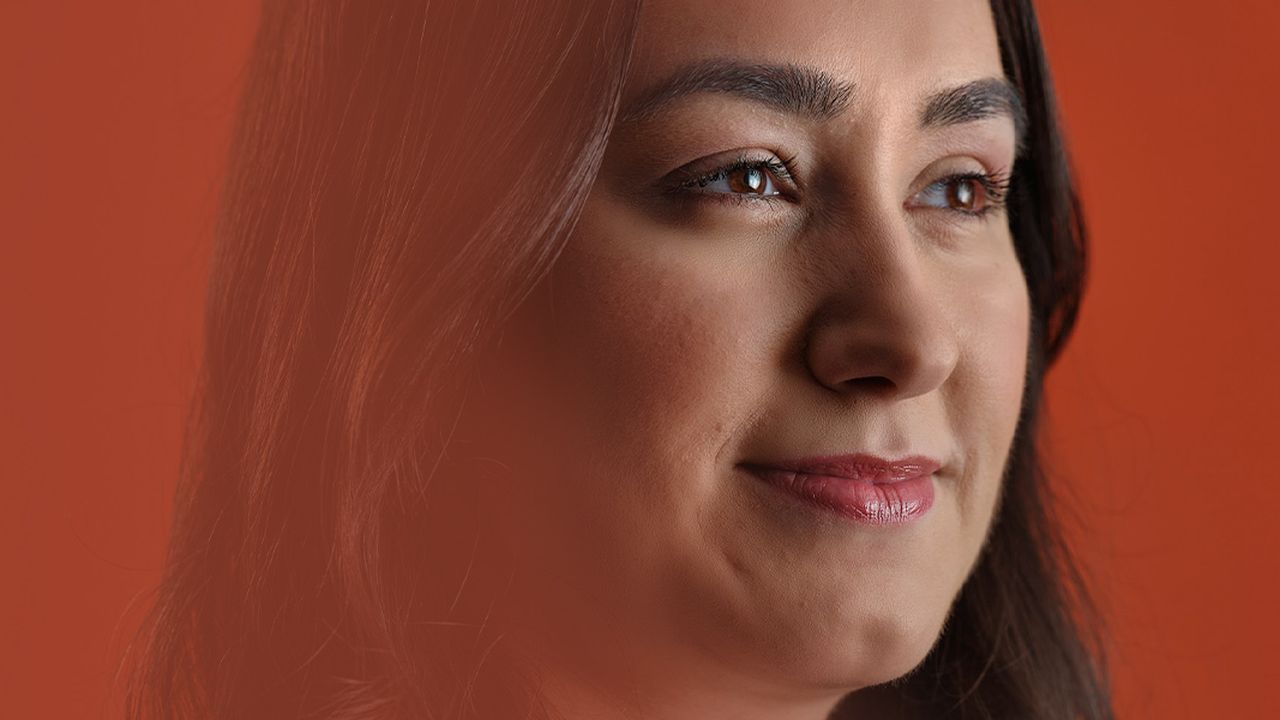 Portrait of a woman with long dark hair against an orange background, gazing thoughtfully to the side.