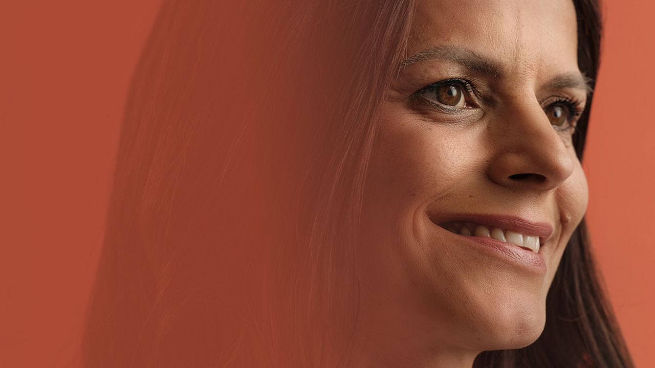 Portrait of a woman smiling, with long dark hair and brown eyes, set against a solid orange background.