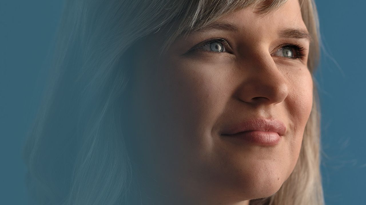 Portrait of a woman with light hair, smiling gently and looking upward against a blue background.