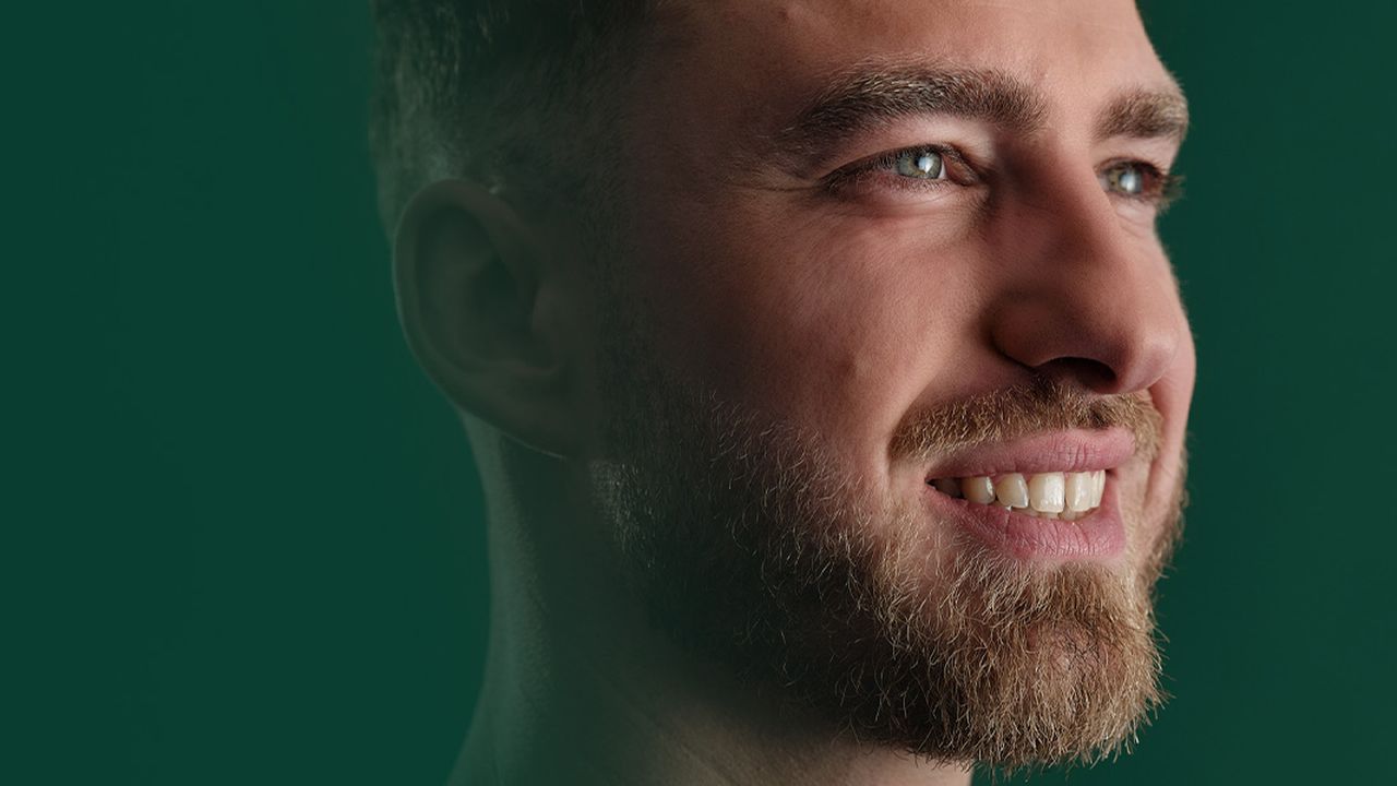 A man with a beard looks to the side, smiling slightly in front of a green background.