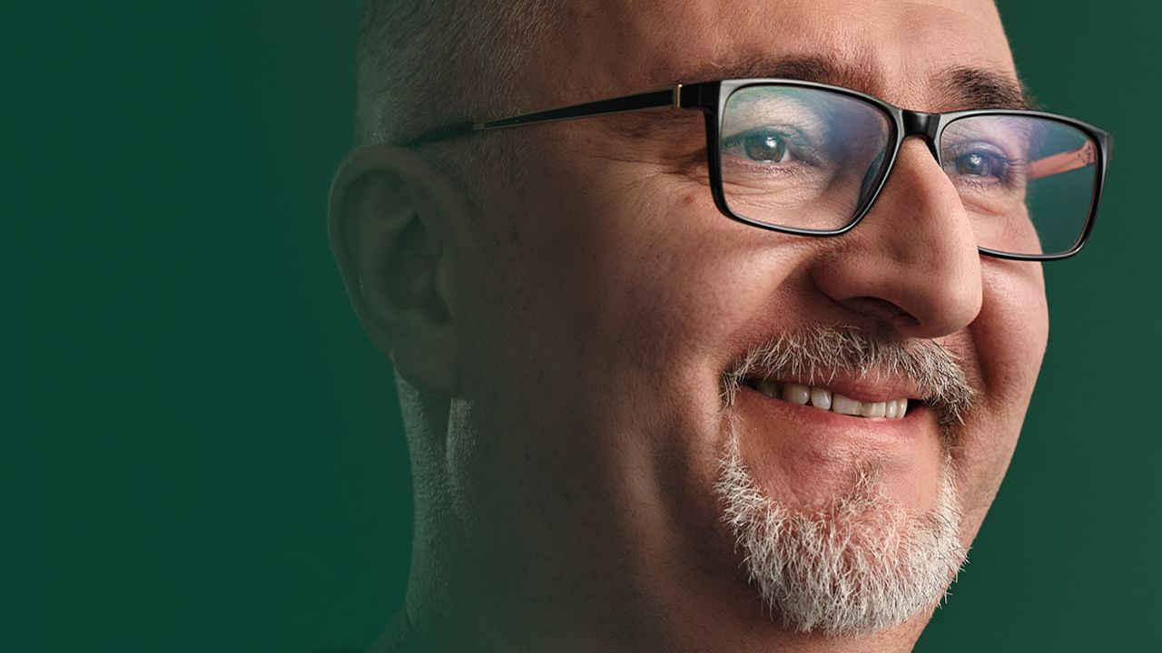 Der Mitarbeiter Nikola steht vor einem grünen Hintergrund und lächelt. Er trägt eine Brille, einen kurzen Bart und einen dunklen Pullover.