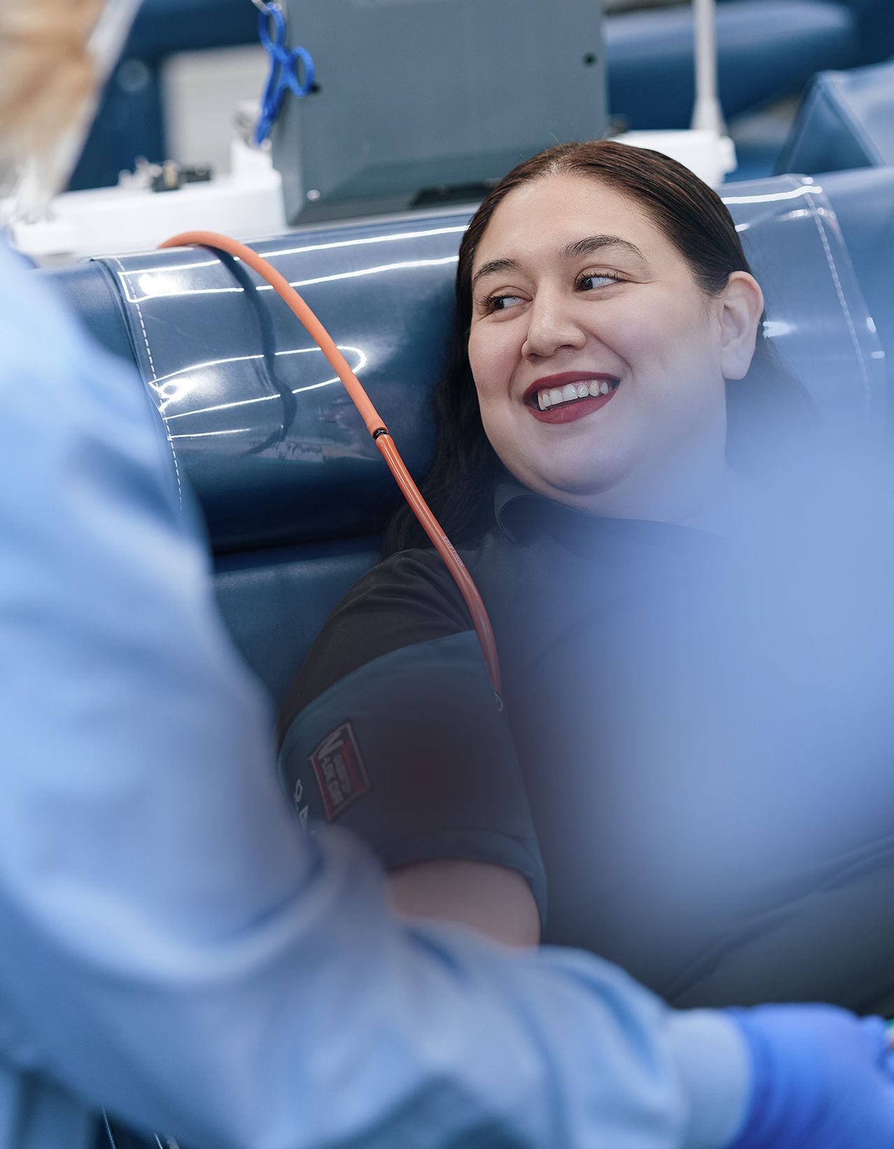 Person smiling while donating plasma, sitting in a medical chair. An attendant in blue scrubs is visible in the foreground.
