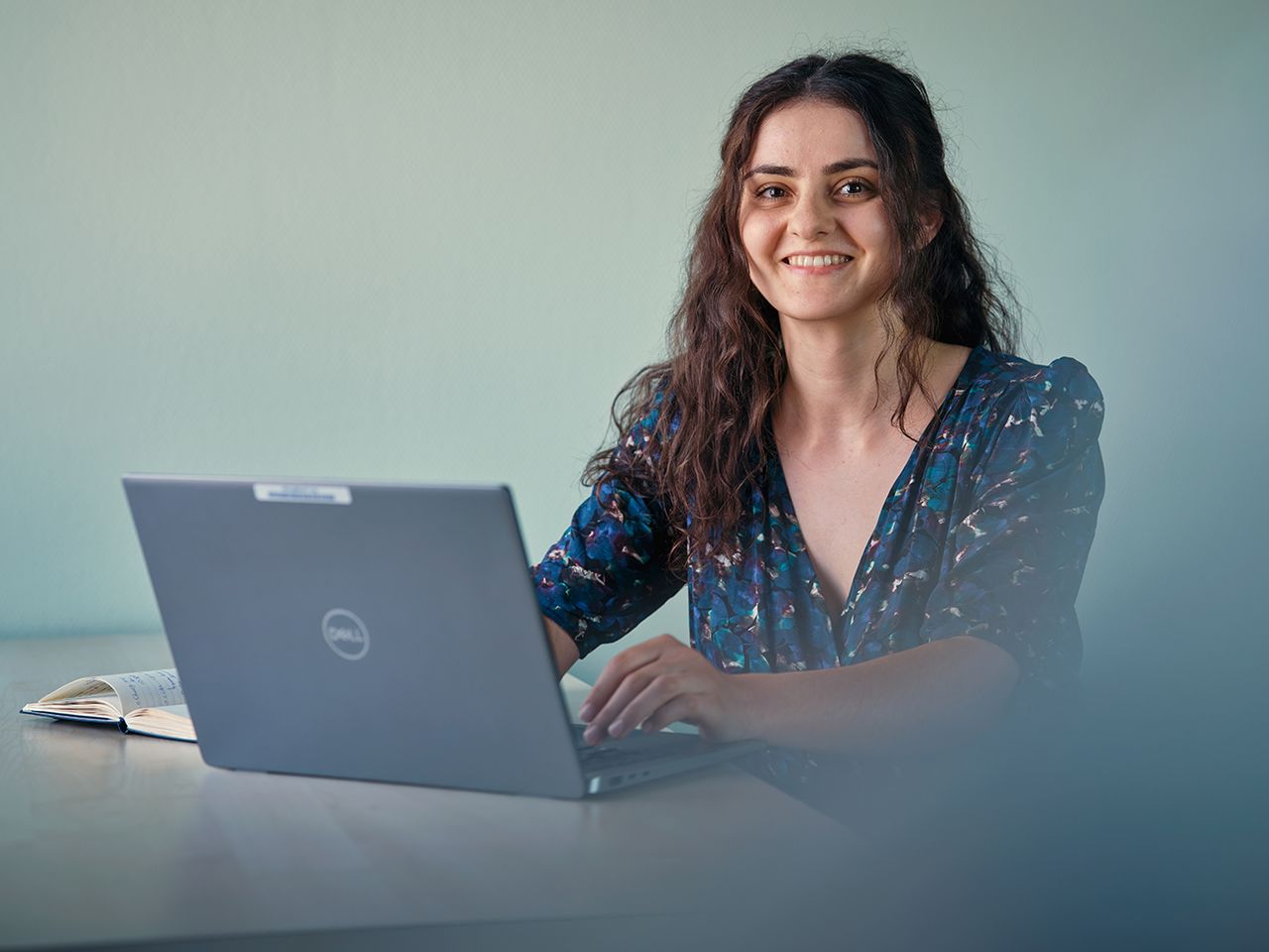 Portrait of Pouneh working on a laptop at a desk, with a notebook beside her. 