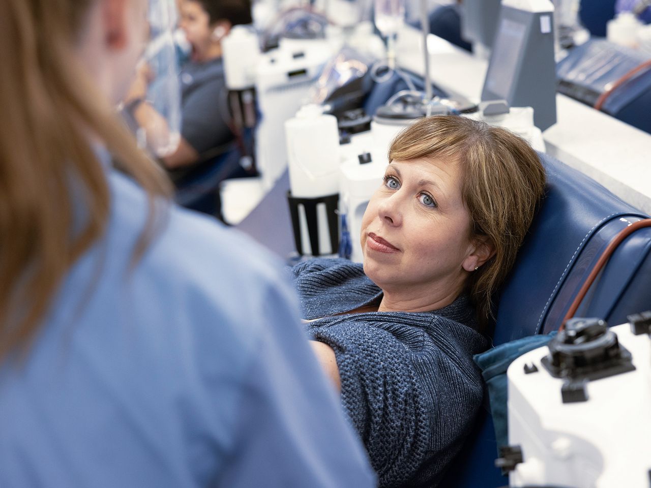 A woman donates blood at a clinic, reclining in a chair and looking at a nurse. Medical equipment is visible in the background.