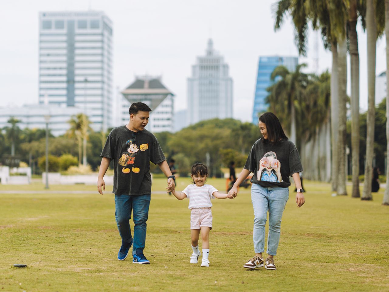 A family of three, holding hands, walks on a grassy area with tall buildings and palm trees in the background.