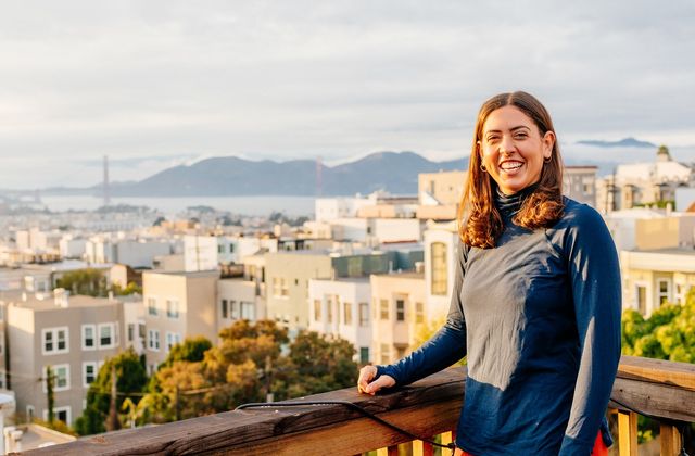 VWD-Patientin lächelt auf einem Balkon mit Blick auf eine Stadtlandschaft mit fernen Bergen und einer Brücke unter bewölktem Himmel.