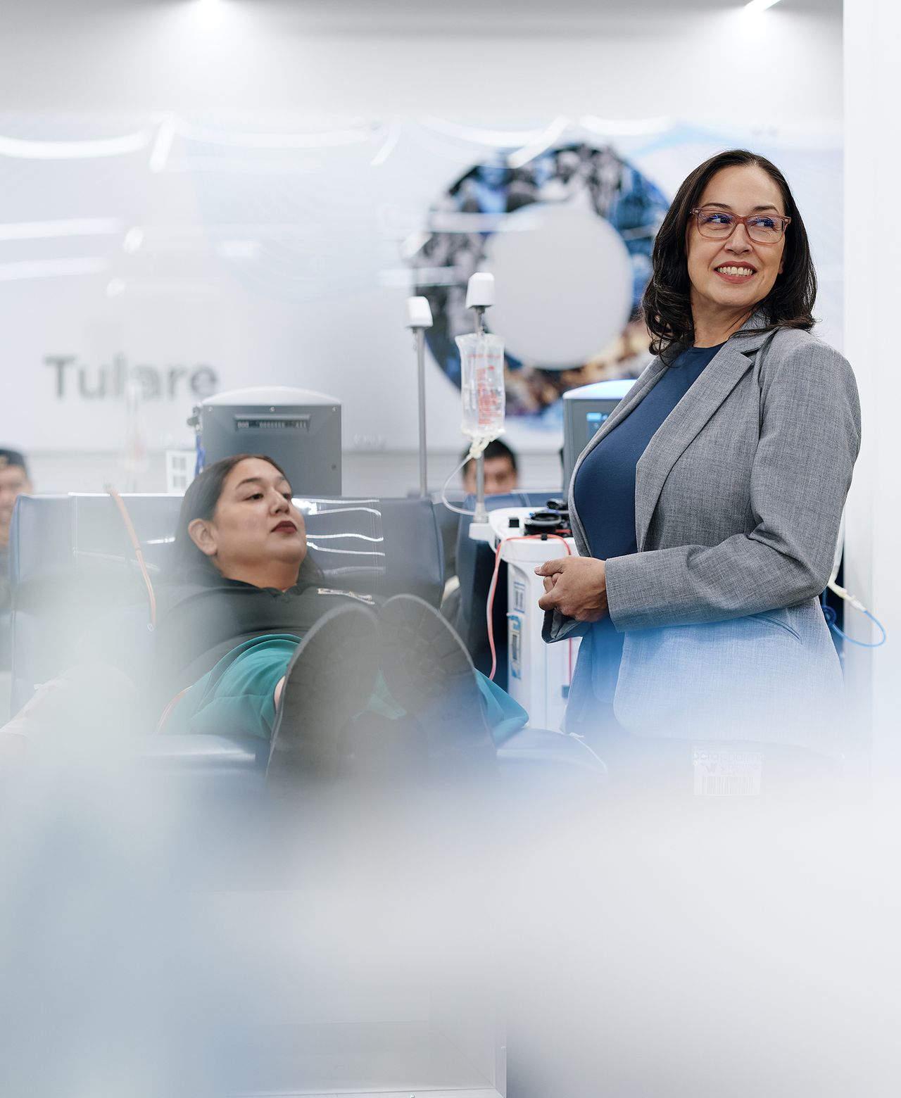 A woman in a gray blazer stands smiling beside a woman donating plasma in a plasma donation centre.